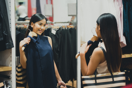 Woman shopping choosing dresses looking in mirror uncertain. Beautiful young asian shopper in clothing store.の写真素材