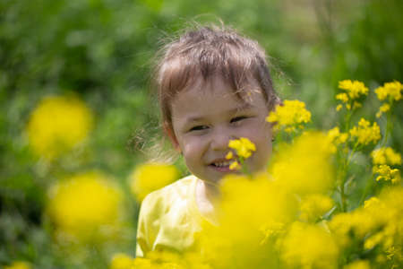 Child girl of mixed race dressed in a yellow T-shirt stands in the sun next to bright yellow flowers...