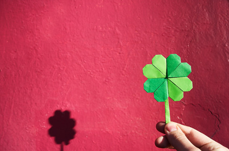 Hand holding paper origami green shamrock on pink wall background.