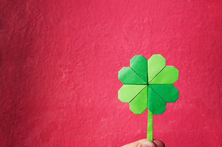 Hand holding paper origami green shamrock on pink wall background.