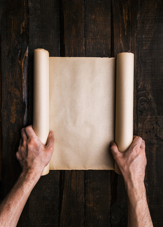 Man hands holding old brown paper scroll on dark wooden planks background. Travel adventure creative...