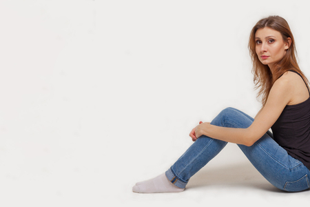 Attractive young caucasian woman with dark brown hair and eyes sitting back to the left side of picture, holding knees with a hands; studio, white background, isolated, copy space.の写真素材