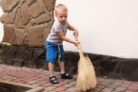 Pretty caucasian baby boy with blonde hair sweeps the yard with a broom. Outdoors, daylight.の写真素材