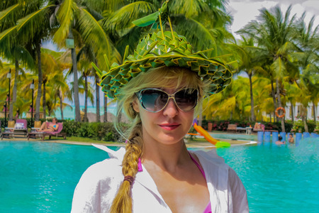 Young attractive woman in sunglasses stands in front of turqueos swimming pool, with the strange hat made from green leaves, with figure of insect (grasshopper) on the top of hat. Outdoors, summer, light sun, copy space. Happy vacation concept.の写真素材