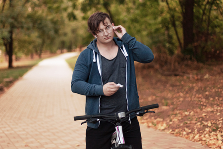 Attractive young caucasian man with dark hair bicycling in the park. Touches the white earphone (airpod). Outdoors, autumn day. Diversity people. Melancholy mood. Round golden glasses, silver ring on one finger, white and pink bicycle. Copy space.の写真素材