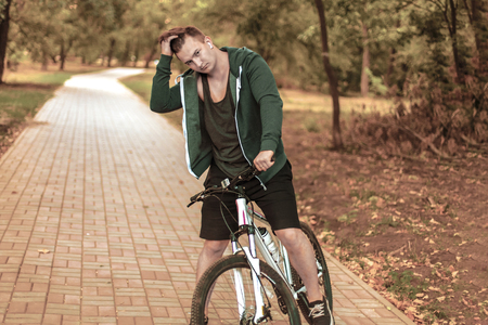 Portrait of romantic young caucasian handsome male with dark curly hair sitting on bicycle alone on abandoned park path. White earphones, earring, casual sportswear, smart seriours look. Healthy life concept, outdoors, copy space. Diversity peopleの写真素材