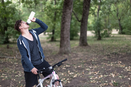Attractive young caucasian man with dark hair drinks water from sport bin, bicycling in the park. Outdoors, autumn/fall park background. Headphones (earphones, airpods), golden round glasses, white and rose bicycle, blue casual sportswear. Copy spaceの写真素材