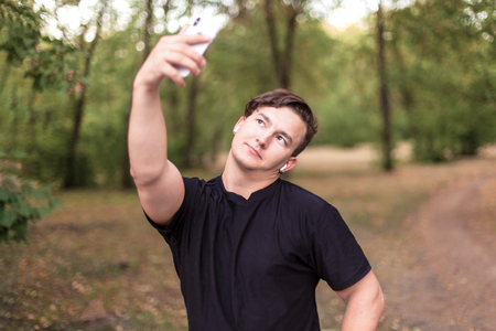 Close up portrait of attractive young caucasian man with dark hair making selfie on mobile phone in the park. Outdoors, pretty autumn day background. Copy space. Black casual, white earphones and earring. Narcissism, diversity people.の写真素材