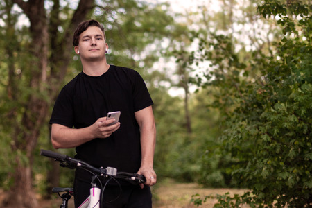 Attractive young caucasian man holds mobile phone and bicycle. White earphones, earring, dark casual wear, dark hair and eyes. Stands at the abandoned park. Outdoors, close up, copy space.の写真素材