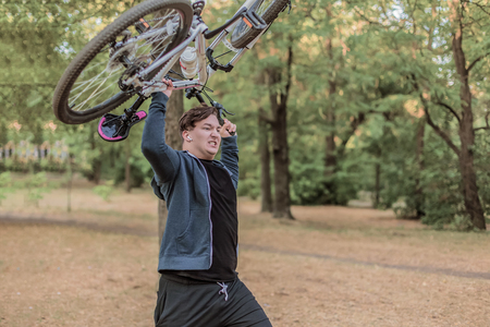 Young emotional cuacasian man crashes the bicycle holdlnig it up in both hands, angry expression on the face. Outdoors, autumn park background. Copy space. Funny picture, humor, concept. Crazy fitness, insurance for health, damned healthy lifestyle.の写真素材