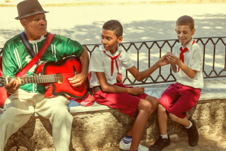 Havana / Cuba 10 Sept 2018: Old musician plays guitar sitting near to two cuban pupils - boys in red and white uniform, smiling to each other. Outdoors, sunny day.のeditorial素材