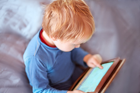 Little caucasian baby boy sits on the sofa using a tablet, touching screen. Red hair, casual wear, indoors, close up, copy space. Children time spending, computerization of youngsters concept.の写真素材