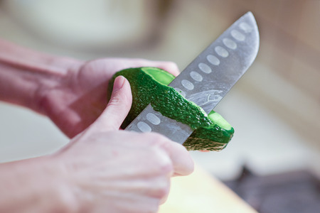Female hands peel fresh organic avocado with knife in kitchen. Close up, indoors, copy space.の写真素材