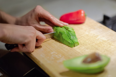 Female hands cutting fresh organic avocado with knife on wooden board in kitchen. Red bell pepper on background. Close up, indoors, copy space.の写真素材