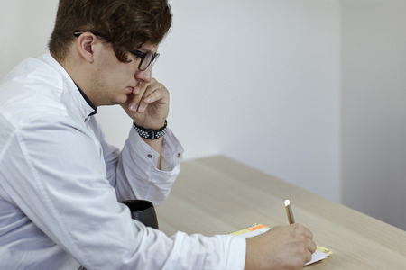 Young caucasian male doctor in a white outfit writes something sitting on the table with cup of tea...