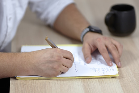 The man' hands on white outfit writes with roller pen on the paper on wooden table (some latin, or medicine terms). The cup of tea of coffee on the table. Student, doctor. Indoors, copy space.の写真素材