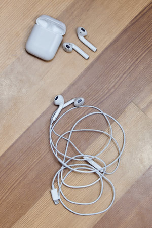 Rostov-on-Don, Russia - Dec. 2018: Apple airpods and wire earphones on wooden blurred background. Apple wireless earphones with charger box, chosen focus. Indoors, copy space, close up.のeditorial素材