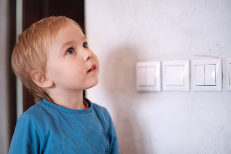 Pretty blonde caucasian baby boy looks up near to the wall with light-switch. Big blue eyes,...