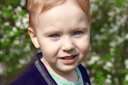 Cute blond baby boy with bright blue eyes smiles in the blossom park. Emotion of happiness, fun, joy. Outdoors, copy space, green and white spring background.の写真素材