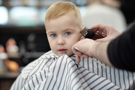 Serious cute blond baby boy with blue eyes in a barber shop having haircut by hairdresser. Hands of stylist with tools (trimmer, brush). Children fashion in salon. Indoors, dark background, copy space.の写真素材