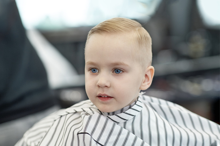 Cute blond smiling baby boy with blue eyes in a barber shop after haircut by hairdresser. Children fashion in salon. Indoors, dark background, copy space.