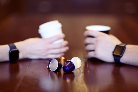 Two hands with equal black electronic wrist watches, holding cups of coffee, white and black, on the...