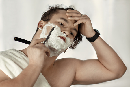 Young handsome caucasian man with electronic watch on wrist shaves with straight razor (vintage style of old barbership). Thoughtful serious look, towel on the shoulder, young metrosexual cares himself. White background, indoors, copy space.の写真素材