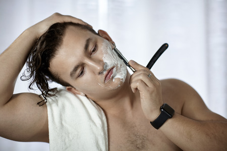 Young handsome caucasian man with electronic watch on wrist shaves with straight razor (vintage style of old barbership). Thoughtful serious look, towel on the shoulder, young metrosexual cares himself. White background, indoors, copy space.の写真素材