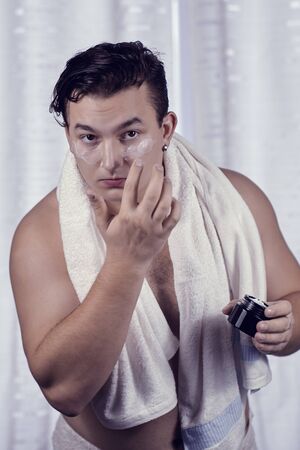 Young handsome caucasian man applying cream under the eyes. Caring face, metrosexual daily routine in the bathroom. Indoors, copy space.の写真素材