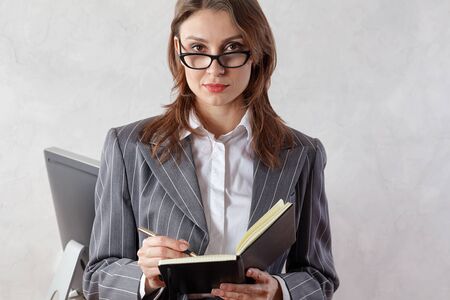 Beautiful young professional brunette woman in office with eyeglasses, writing in a pad, with confident expression. Classic business style (striped suit). Indoors, light backgroundの写真素材