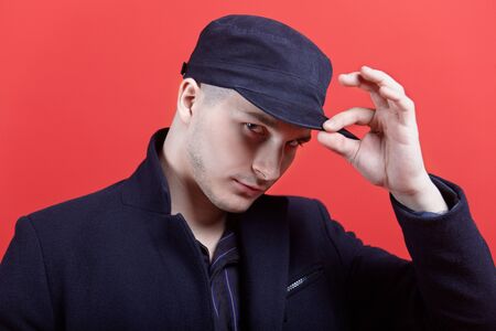 The portrain of young handsome caucasian man touching the black cap. Total black wear, expressive look at camera. Indoors, red background, copy space, logo presentation.の写真素材