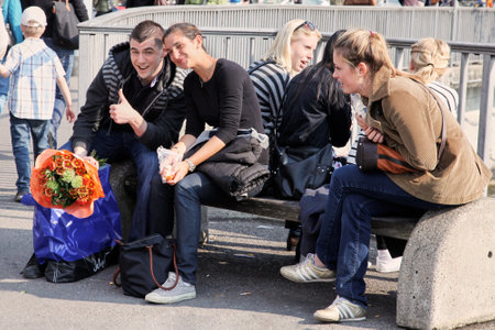 Geneva, Switzerland - May, 2012: The group of young people sitting on the street bench in front of the river. Couple of friend, man with a bouquet and thumb up, woman smiling. Youngsters on the street, outdoors.のeditorial素材