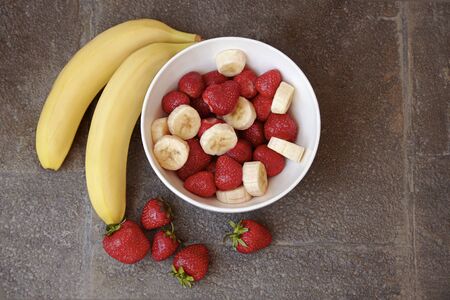 Healthy meal with strawberry and bananas. Organic natural diet concept, real natural fruits, fresh and washed, cut on pieces in  white bowl on dark stone surface. Copy space.の写真素材