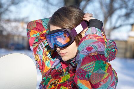 Young attractive woman in bright sportswear puts on special glasses for snowboarding and winter sport. Outdoors, snowy  background. Active and positive lifestyle.の写真素材