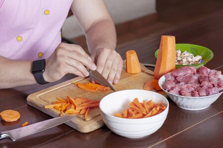 Man hands cutting vegetables for cooking. Carrot and mushrooms, row meat, ingredients for stew or ragout. Classical brown wooden kitchen interior, pink shirt, black watch on the wrist, modern male lifestyle, hobby or routine. Indoors.の写真素材