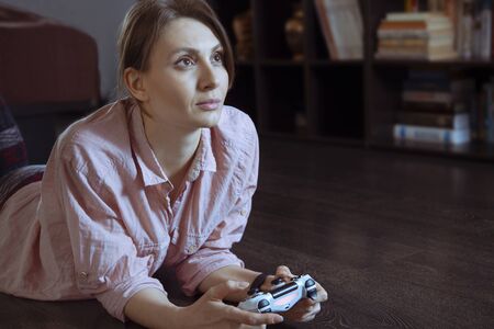 Young attractive woman with dark hair and eyes plays computer game, laying on the floor of her room....