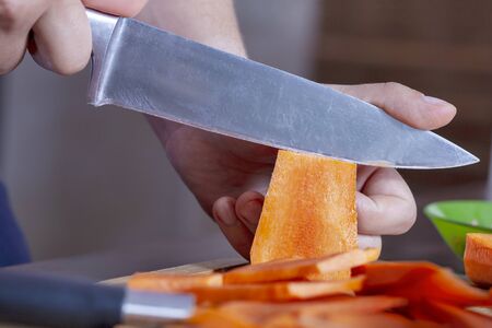 Close up man hands cutting row carrot for cooking with big knife, on large pieces. Ingredients of vegetable cuisine or natural diet. Hobby or daily routine of modern man. Indoors.の写真素材