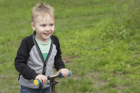 Cute blond caucasian boy with blue eyes having fun at park with kick scooter. Pretty smiling baby face, positive emotions, active time spending, happy childhood. Outdoors, copy space.の写真素材