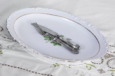 Old vintage silver knife and fork set on empty oval plate on the table covered with white embroidered tablecloth. Grandparents or haute cuisine style, luxury or dieting concept. Indoors, copy space.の写真素材
