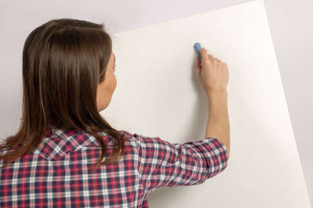 Young beautiful woman with dark brown hair writes something with bright blue chalk on white desk....