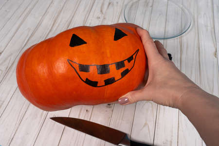 Human hand holding Halloween pumpkin with drawing Jack-o-lantern face, kitchen knife is near on the table. Copy space, preparing holiday concept.の写真素材