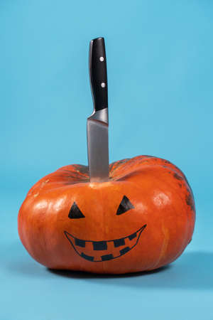 Halloween pumpkin with drawing Jack-o-lantern face and kitchen knife sticking out from above, on blue background. Copy space, holiday concept.の写真素材