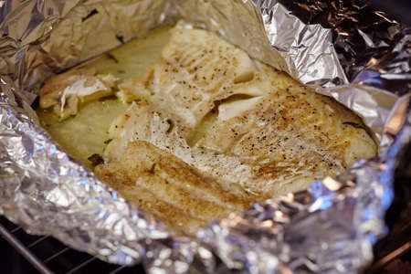 Close up fillet of white fish in the foil, sprinkled with black pepper and aromatic herbs, on a baking sheet removed from the oven. Healthy diet concept. Soft selective focus.の写真素材
