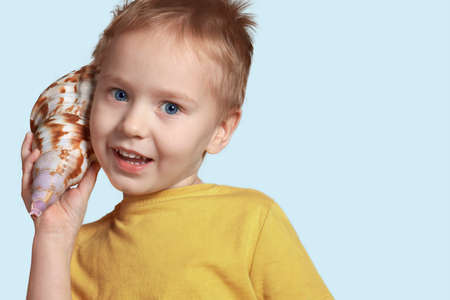 Little cute caucasian boy with blond hair and blue eyes listens to seashell, smiling. Pretty baby, indoors, copy space, isolated.の写真素材