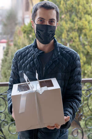 Serious middle-aged man stands in front of the door holding cardboard box with bow, gift for friend. Spring time, black mask on the face. Outdoors, copy space.の写真素材