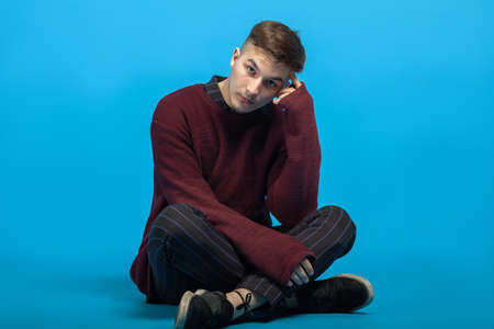 Young handsome man sits on the floor on blue background. Fashion hairstyle, expressive look right to...