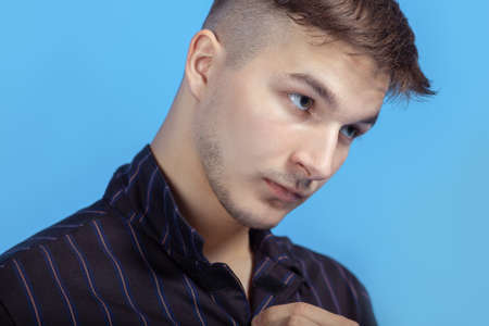 Closeup portrait of young handsome man with serious glance, unbuttoning the shirt. Trendy hairstyle, short accurate beard, blue background. Sensual look. Copy space.の写真素材