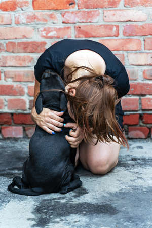 Young caucasian woman embraÑes little black puppy of Staffordshire Bullterrier breed. Cute picture on red brick wall background, friendship of human and dog. Outdoors, copy space.の写真素材