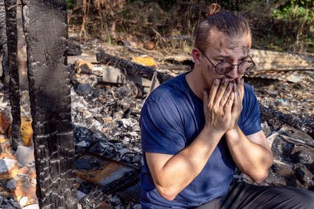 Young man sits on the ruins of house destroyed with fire. Damaged window, walls and floor, burnt wooden frames. Tragic face, strong emotions, crying. Insurance or emergency concept, misadventure, disaster.の写真素材