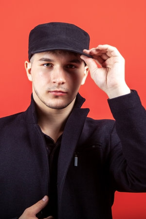 Portrait of young handsome caucasian man touching his black cap with confident look right to the camera. Formal wear, emotional face expression. Red background, copy space.の写真素材